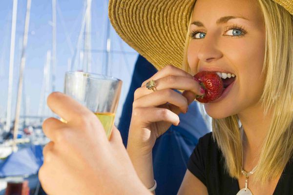 زن جوان بلوند فوق العاده زیبا و ثروتمند در عرشه قایق بادبانی خود در یک مارینا نشسته و توت فرنگی می خورد و شامپاین می نوشد