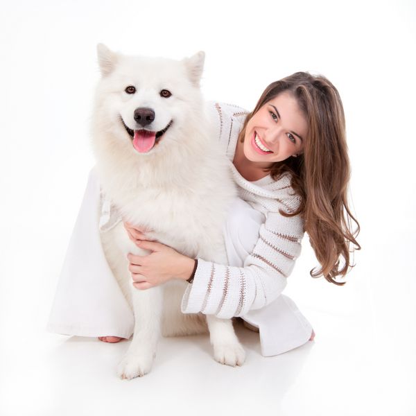 تصویری استودیویی از زن جوانی که لباس سفید پوشیده سگ سفیدش را در آغوش گرفته است هر دو ژست گرفته اند شاد و خندان به نظر می رسند