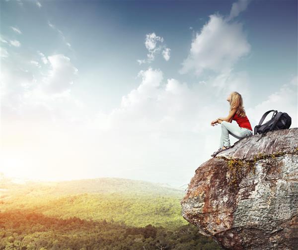 زن جوان با کوله پشتی که روی لبه صخره نشسته و به آسمانی با ابرها نگاه می کند