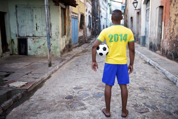 بازیکن فوتبال برزیلی با پیراهن سال 2014 در حالی که یک توپ فوتبال در یک خیابان قدیمی فاولا در دهکده روستایی در دست دارد ایستاده است