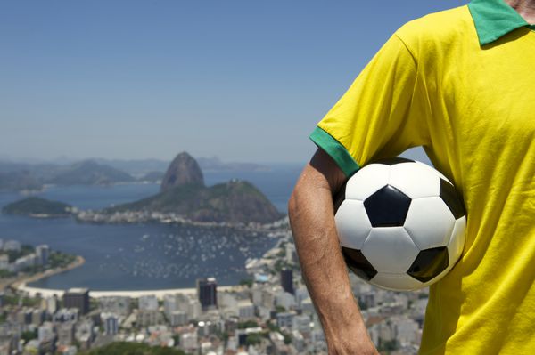 فوتبالیست برزیلی با پیراهن به رنگ برزیل در افق ریودوژانیرو با کوه شوگرلوف فوتبال در دست دارد