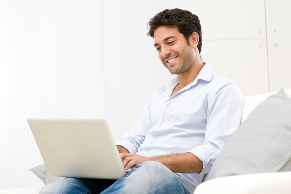 مرد جوان خندان خوشحال در حال تماشا و کار با لپ تاپ کامپیوتر در خانه