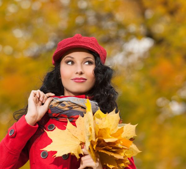 زن جوان قفقازی جذاب با لباس های رنگارنگ گرم روی برگ های زرد در فضای باز و خندان
