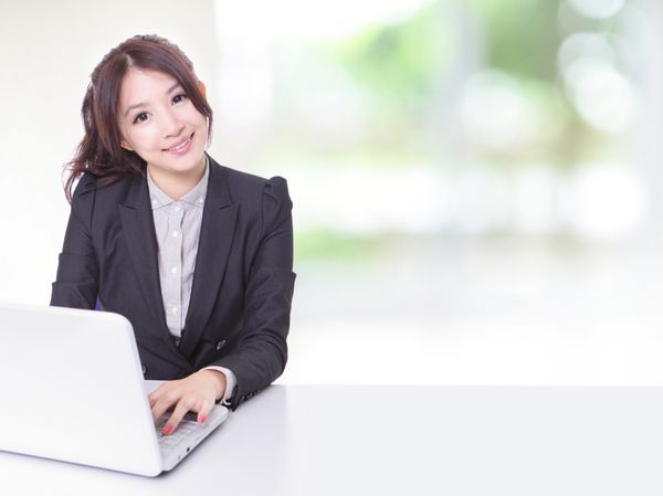 زن تجاری جوان با استفاده از کامپیوتر و نشستن در دفتر شرکت با میز سفید پنجره بیرون پس زمینه سبز است مدل یک زیبایی آسیایی است