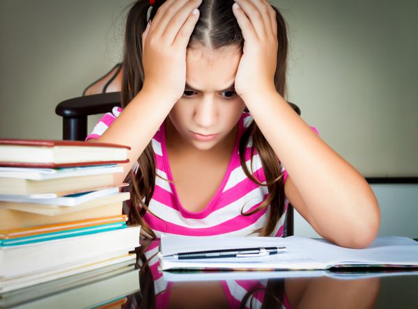 دختر مدرسه ای عصبانی و خسته در حال مطالعه با انبوهی از کتاب ها روی میزش