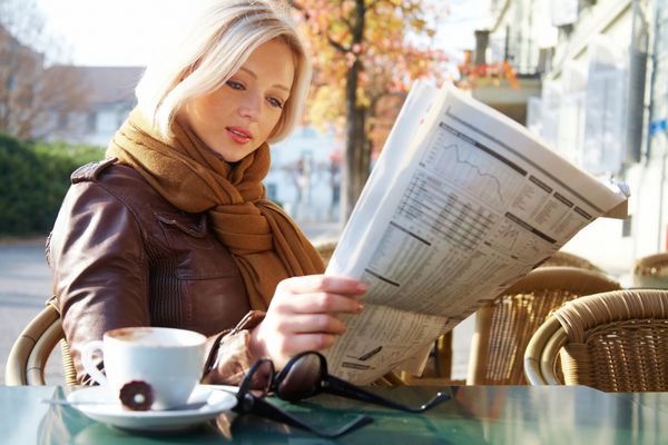 زن تاجر جوان جذاب در حال خواندن روزنامه در کافه در فضای باز