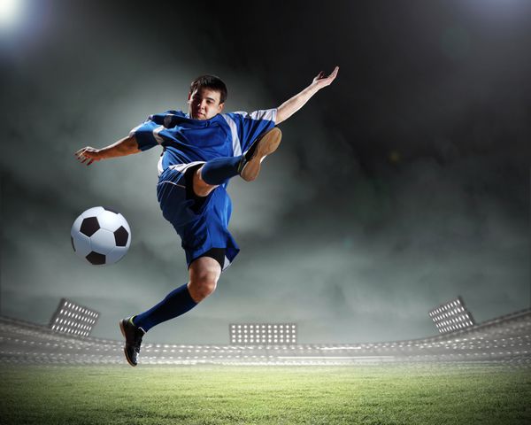 بازیکن فوتبال با پیراهن آبی در حال ضربه زدن به توپ در ورزشگاه