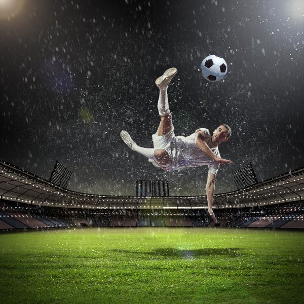بازیکن فوتبال با پیراهن سفید در حال ضربه زدن به توپ در استادیوم زیر باران