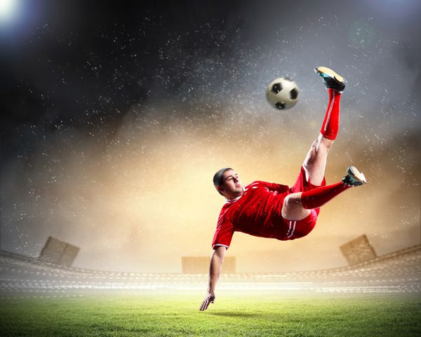 بازیکن فوتبال با پیراهن قرمز در حال ضربه زدن به توپ در استادیوم زیر باران