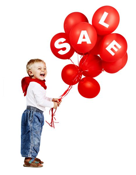 پسر کوچولوی ناز با پیراهن سفید شلوار جین و روسری قرمز در حالی که یک دسته بادکنک قرمز با علامت فروش در دست دارد می خندد