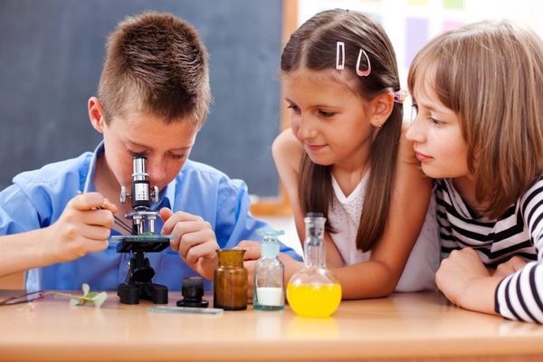 پسر برجسته مدرسه ابتدایی در حالی که دختران در حال تماشای آن هستند به میکروسکوپ نگاه می کند