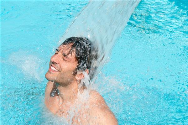 مرد جوان شادی که در حال تفریح و استراحت زیر یک جت آب در استخر شنا است
