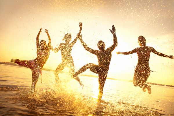 گروهی از جوانان شاد در حال رقصیدن و سمپاشی در ساحل در غروب زیبای تابستان