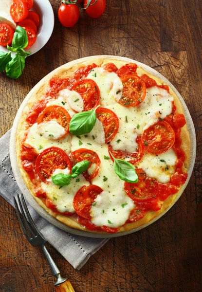 نمای بالای یک پیتزای رنگارنگ گوجه فرنگی و پنیر تازه پخته شده با گیاهان تازه و برگ های ریحان پاشیده شده و در بشقاب روی میز چوبی قدیمی سرو می شود