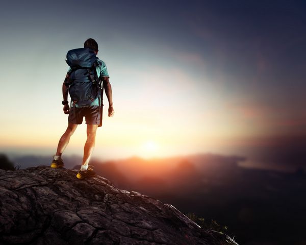 کوهنورد با کوله پشتی که در بالای کوه ایستاده و از طلوع خورشید لذت می برد