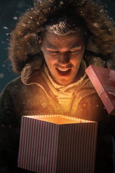 مرد جوان خوشحالی که در شب کریسمس جعبه هدیه را در فضای باز باز می کند