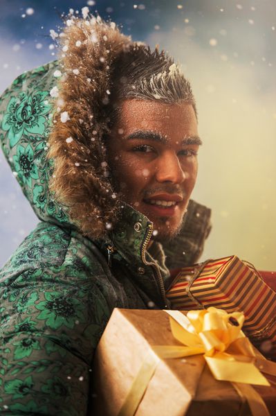 تصویر روشن مرد خوش تیپ در حال حمل هدایای کریسمس در فضای باز در زمستان در شب نور طلایی و زرق و برق جادویی در اطراف او