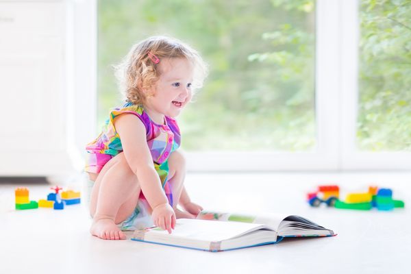 دختر بچه نوپا با موهای مجعد بلوند در حال خواندن کتاب روی زمین نشسته و در یک اتاق خواب سفید با پنجره های بزرگ و با اسباب بازی های اطرافش کتاب می خواند