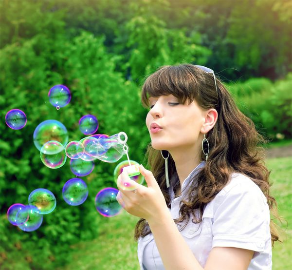 دختر جوان رمانتیک در حال باد کردن حباب های رنگارنگ صابون در پارک بهار