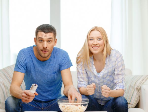 غذا عشق خانواده ورزش سرگرمی و مفهوم شادی - زوج خندان با پاپ کورن در حال تشویق تیم ورزشی در خانه