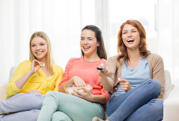 مفهوم خانه فناوری و دوستی - سه دختر نوجوان خندان در حال تماشای تلویزیون در خانه و خوردن ذرت بو داده