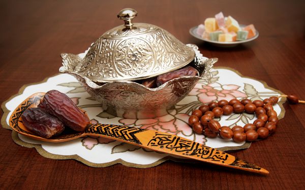 یک کاسه و قاشق چوبی پر از میوه های خرمای خرما که نماد ماه رمضان است و در پس زمینه آن شیرینی های ترکی است عبارت بسم الله حک شده روی قاشق چوبی