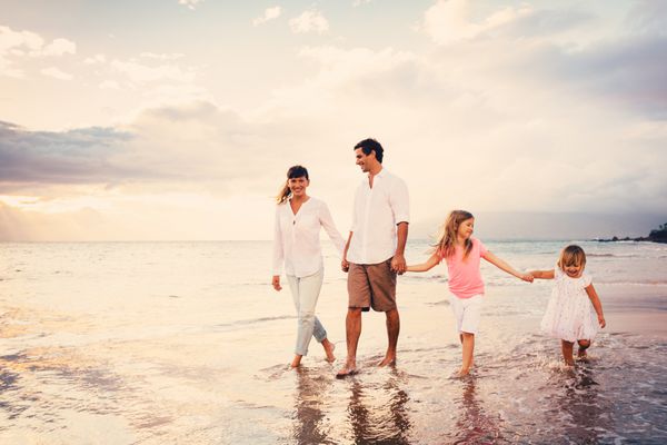 خانواده جوان شادی که با قدم زدن در ساحل در غروب آفتاب سرگرم می شوند