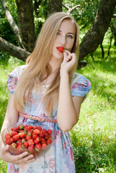 تصویری از دختر نوجوان خوش شانس دختر نوجوان شاد در حال خوردن توت فرنگی از کاسه بزرگ در باغ سبز روز تابستان در پس زمینه در فضای باز