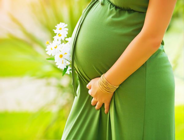نمای نزدیک روی شکم زن باردار لباس سبز بلند پوشیده دسته گل گل های دیزی را در دست در خارج از منزل مفهوم جدید زندگی