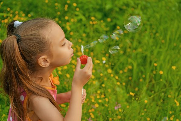 دختر کوچک شاد و زیبا در پارک سبز تابستانی حباب ایجاد می کند