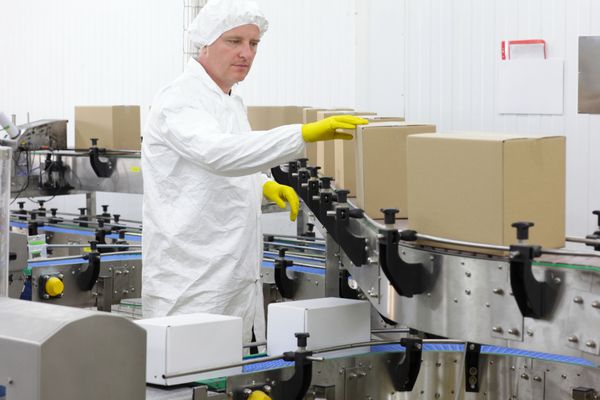 کارگر مرد قفقازی با پیش بند سفید کلاه و دستکش زرد در خط تولید در کارخانه