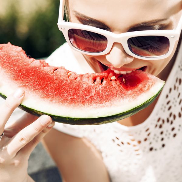 دختر جوان با لباس تابستانی سفید با عینک آفتابی هندوانه آبدار را گاز می گیرد