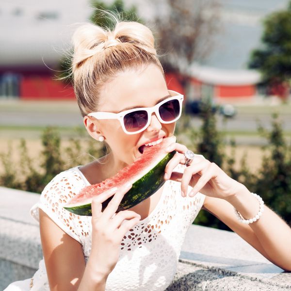 دختر بلوند جوان با لباس تابستانی سفید و عینک آفتابی با هندوانه آبدار گاز می گیرد