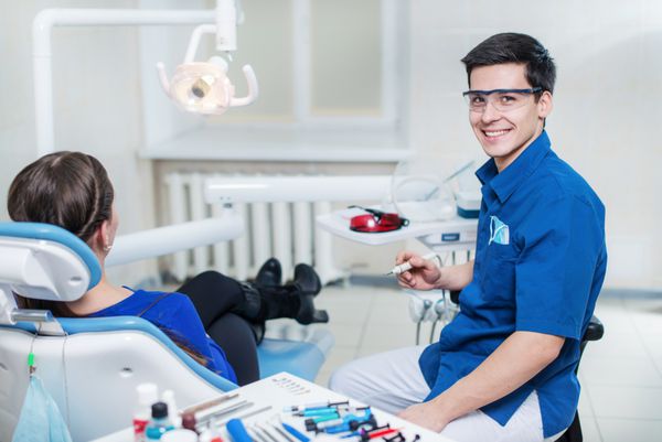 سلامتی در لبخند دندانپزشک موفق و خندان دندان های یک بیمار جوان و دستگاه بور در دست را درمان می کند و مستقیم به دوربین نگاه می کند