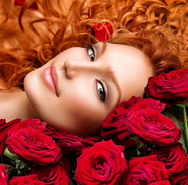 دختر مدل زیبایی با موهای مجعد قرمز بلند و دسته گل رز قرمز زیبا مدل مو با گل زن مد با موهای سالم موج دار که روی رزهای زیبا خوابیده است موی فر شده