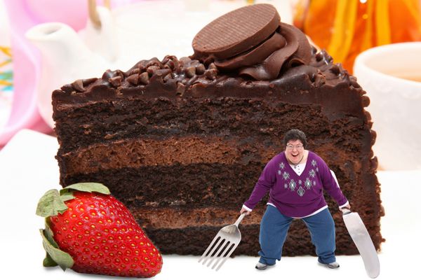 زن قفقازی چهل و پنج ساله هیجان زده با چنگال و چاقو روی بشقاب بزرگ کیک شکلاتی تیره و شیری با توت فرنگی ایستاده است