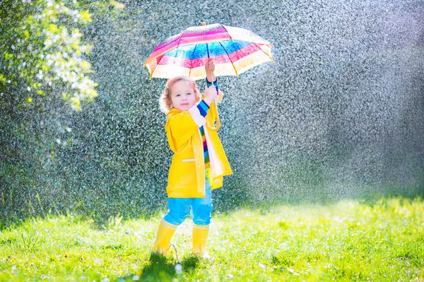 دختر نوپای فرفری بامزه با کت و چکمه های زرد رنگ ضد آب و چتر رنگارنگ در دست دارد و در هوای باران و آفتاب در یک روز گرم پاییزی یا تابستانی در باغ بازی می کند