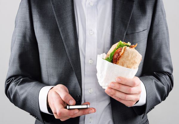 مرد تاجری مشغول در حال حرکت با کت و شلوار با استفاده از تلفن هوشمند در یک دست و نگه داشتن ناهار رول BLT ciabatta در دست دیگر