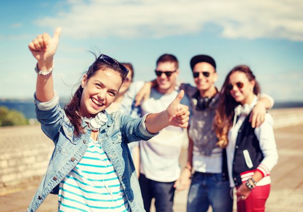 تعطیلات تابستانی و مفهوم نوجوانی - دختر نوجوان با عینک آفتابی و هدفون در حال معاشرت با دوستان در بیرون و نشان دادن شست
