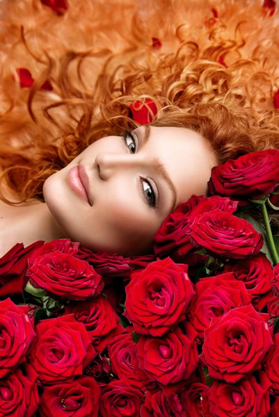 بیوتی مد زن با موهای مجعد قرمز بلند و دسته گل رز قرمز زیبا مدل مو با گل دختر مدل زیبا دراز کشیده روی رزهای زیبا موهای سالم موج دار موی فر شده