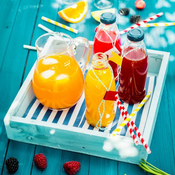 بطری‌های آب پرتقال و توت تازه فشرده شده روی یک سینی چوبی روی میز پیک‌نیک آبی فیروزه‌ای رنگارنگ در آفتاب تابان تابستانی ایستاده‌اند