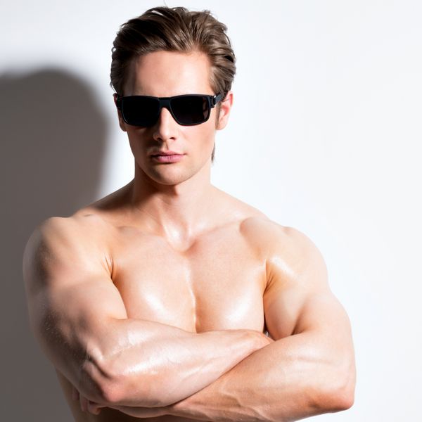 مرد خوش اندام عضلانی با عینک با دستان متقاطع در استودیو به عنوان مدل لباس در زمینه سفید با سایه های متضاد