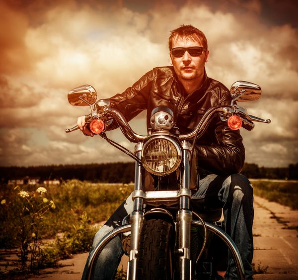 مرد دوچرخه‌سواری با یک ژاکت چرمی و عینک آفتابی که روی موتور سیکلت خود نشسته و به غروب آفتاب نگاه می‌کند