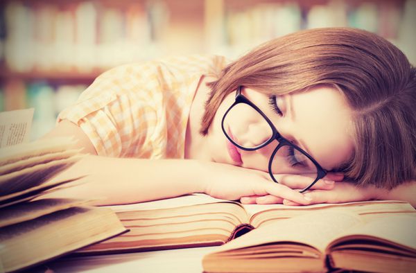 دختر دانشجوی خسته با عینک که روی کتاب های کتابخانه خوابیده است