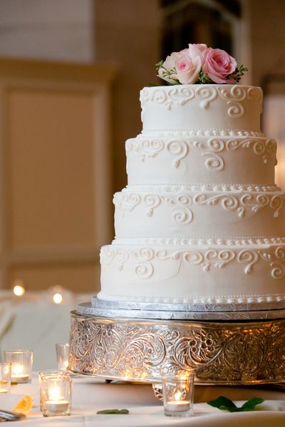 یک کیک عروسی چند سطحی سفید روی پایه نقره ای و گل های صورتی روی آن