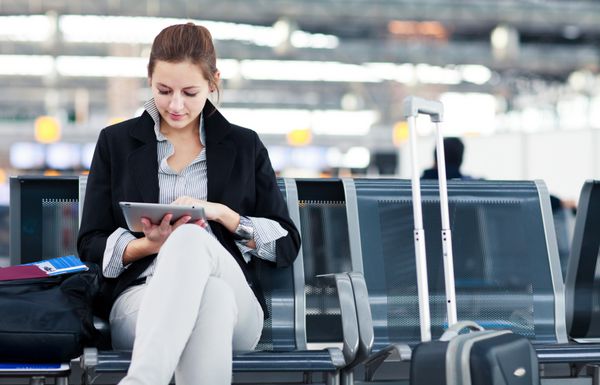 مسافر زن جوان در فرودگاه با استفاده از رایانه لوحی خود در حالی که منتظر پرواز خود است تصویر رنگارنگ