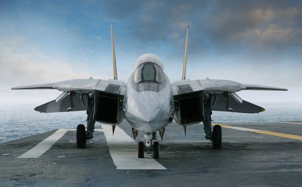 یک جنگنده جت F-14 روی عرشه ناو هواپیمابر در زیر آسمان آبی و ابرها از جلو مشاهده می شود