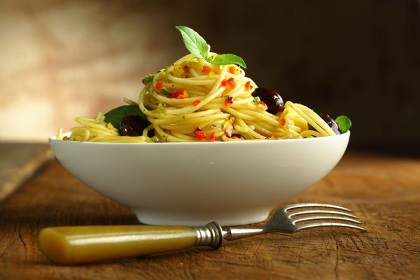 از نزدیک یک کاسه سفید پر از ماکارونی اسپاگتی و زیتون و یک چنگال