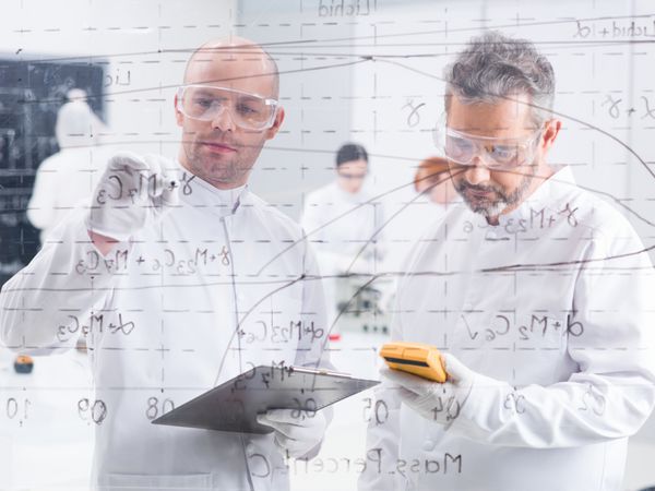 نمای نزدیک از دو محقق در آزمایشگاه که فرمول ها را روی یک تخته شفاف می نویسند و نتایج را با دو دانش آموز دیگر در پس زمینه اسکن می کنند