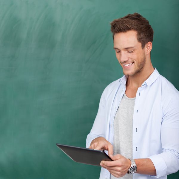 دانشجوی پسر جوان خندان با استفاده از تبلت دیجیتال در برابر تخته سیاه سبز